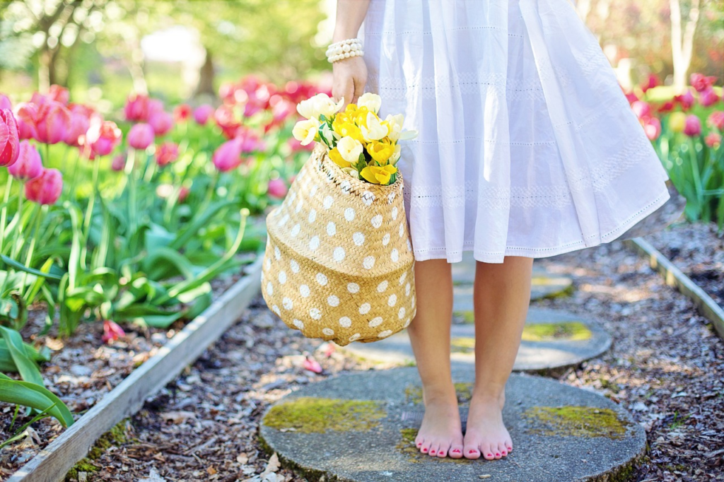 Ein Garten mit blühenden rosa Tulpen. Auf dem Gartenweg steht eine Frau im weißen Sommerkleid barfuß mit pink lackierten Fußnägeln. In der Hand hält sie eine gelbe Basttasche, aus der gelbe Tulpen schauen.